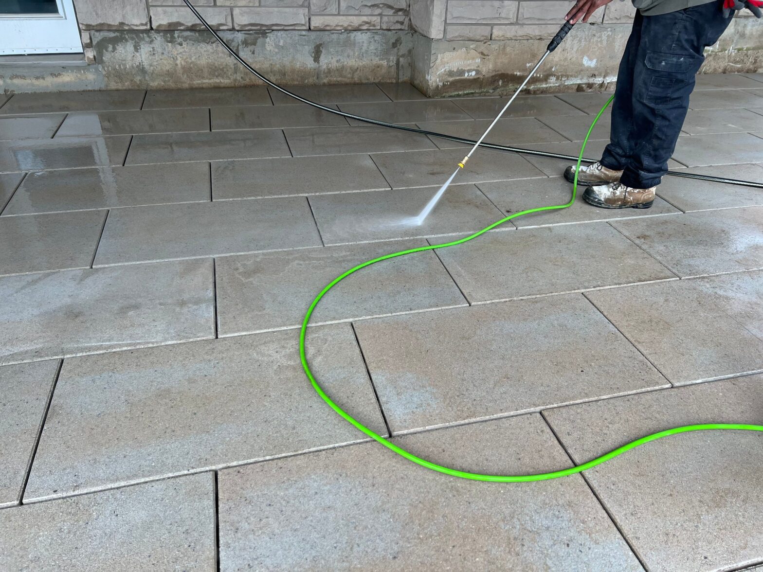 A worker using a pressure washer to clean large interlocking pavers on a patio, with visible water spray and a green hose.