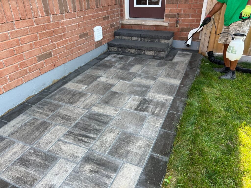 A professional applying a sealant to a newly installed interlocking stone walkway, showcasing clean and vibrant pavers beside a brick house.