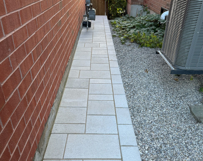 Narrow paver stone walkway consisting of grey rectangular pavers of varying sizes flush against the side of a house, exemplifying Interlocking stone walkways on a smaller walkway